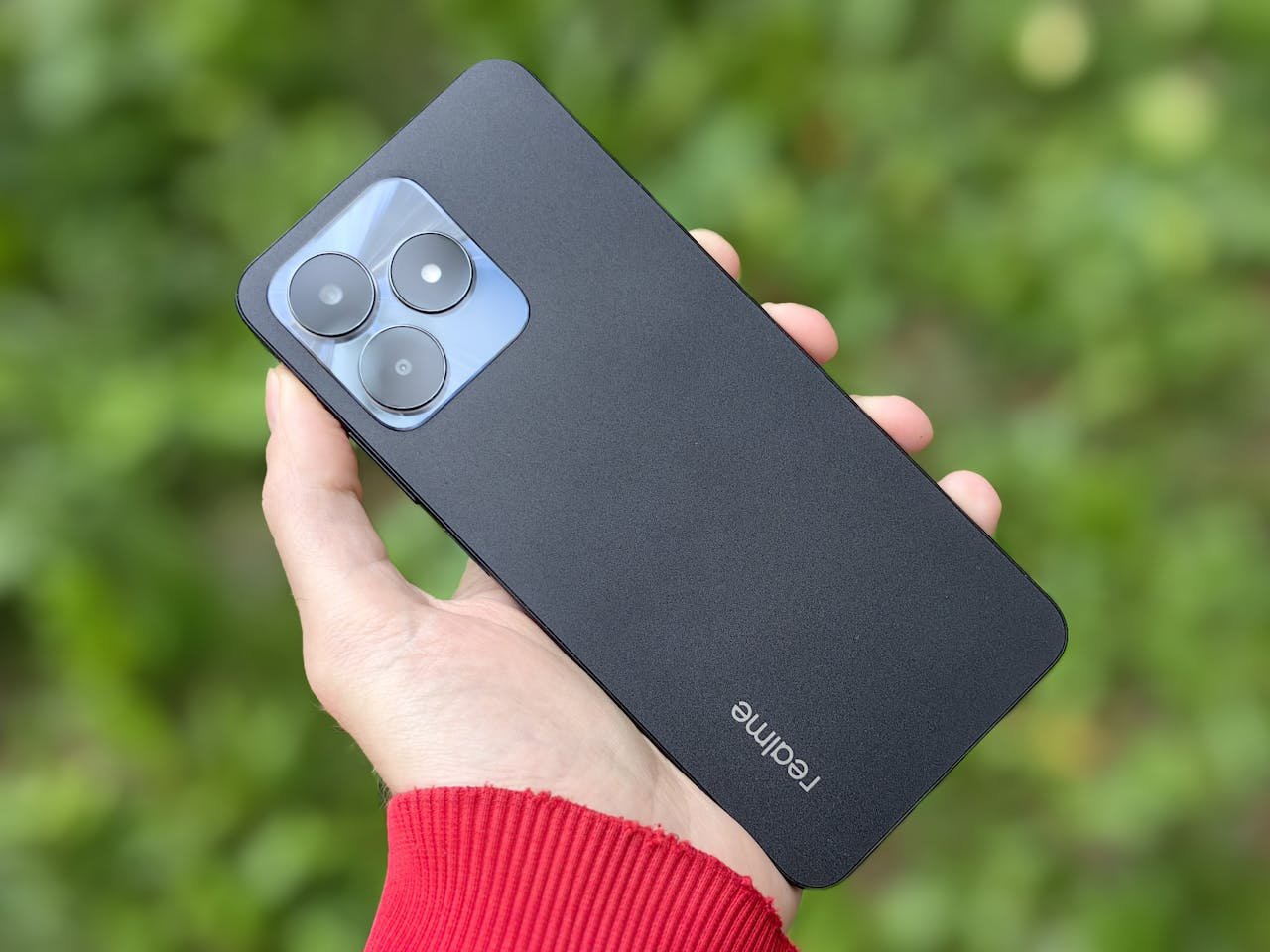 A close-up shot of a hand holding a Realme smartphone with a blurred green background, showcasing modern technology outdoors.